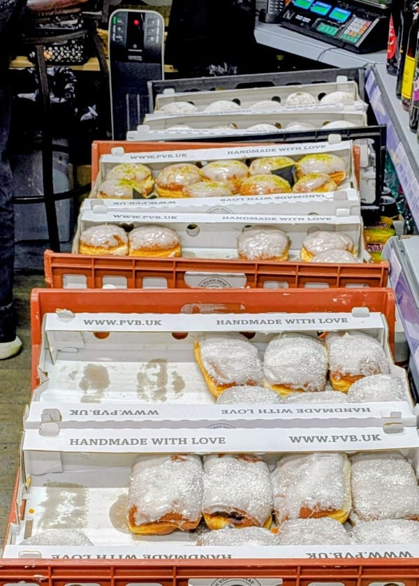 A line of rust-red coloured plastic bakers trays behind the till counter filled with different kinds of doughnuts. The ones in the foreground are the folded cheese doughnuts. The rest are either dusted with sugar or covered in white icing. Some of the iced doughnuts are sprinkled with pistachios