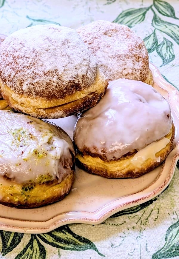 A cream cermaic wavy-edged plate on a tablecloth patterned with green and white lily of the valley leaves and flowers. On the plate, five doughnuts. At the front, an iced pistachio and an iced custard and rosehip jam, on top and behind, plum butter-filled
